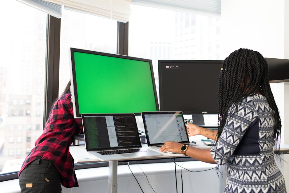 Two women working on laptops and monitors in a bright office setting, focused on technology and teamwork.